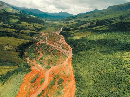 A landscape showing a rust stained river in a green valley with mountains in the background.