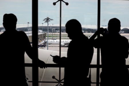 Photograph looking outwards from an airport window to a parked Signapore Airlines flight on the tarmac as passengers seen in silhouette stand in front of the window taking photos, looking at the plane, and using their phones